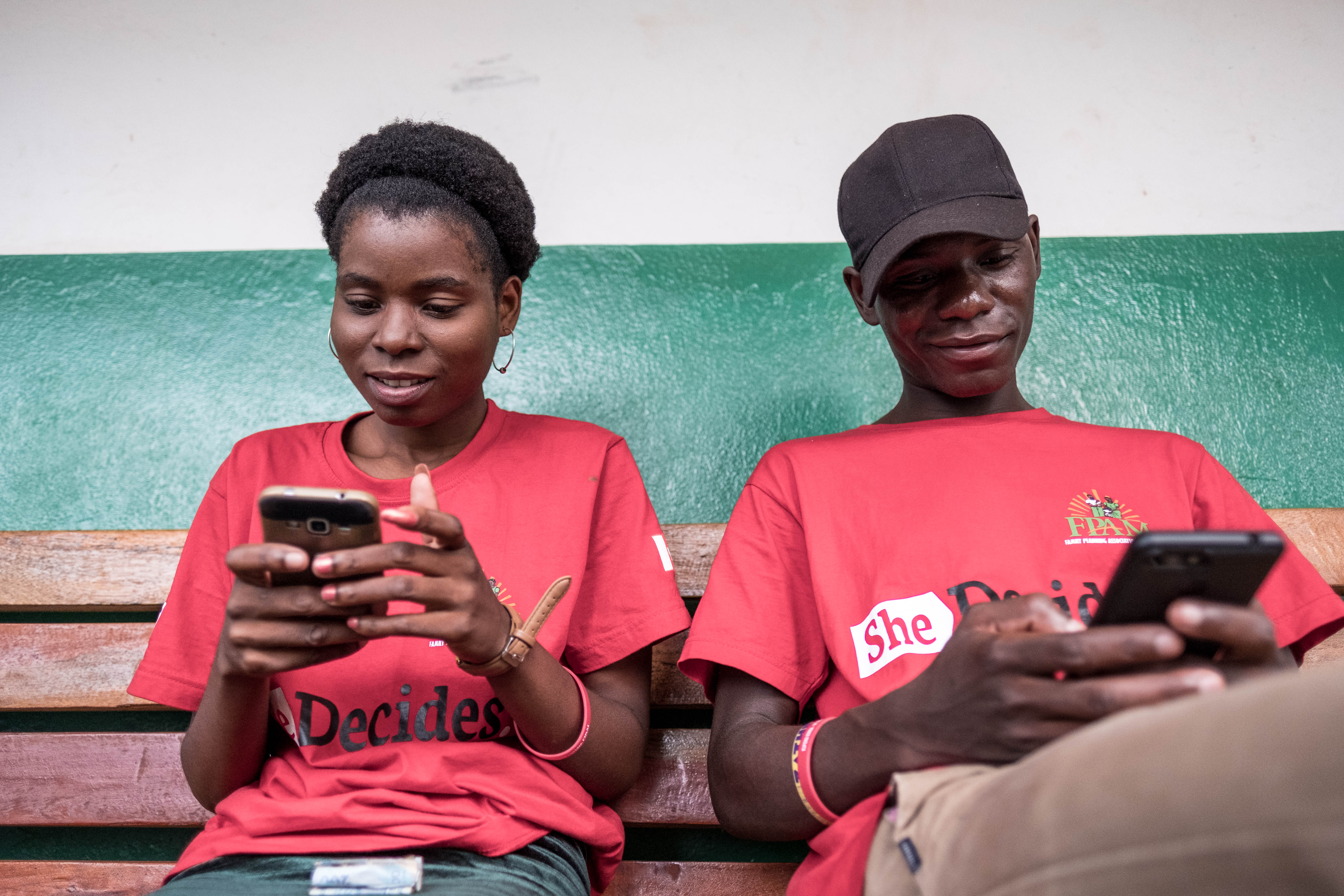The image shows two people in red t-shirts both holding a mobile phone.