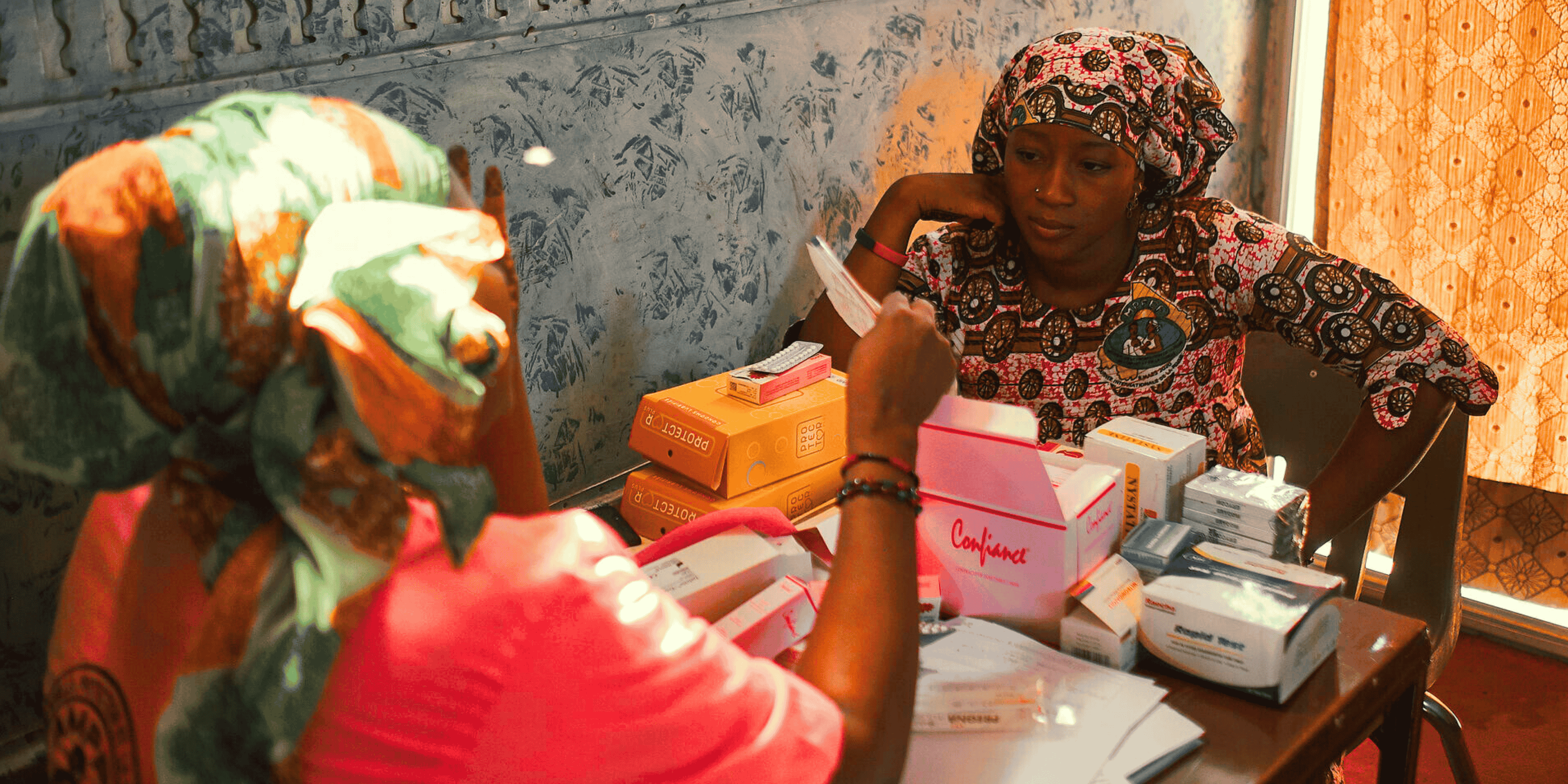 Two women sit at a table with boxes of contraceptives