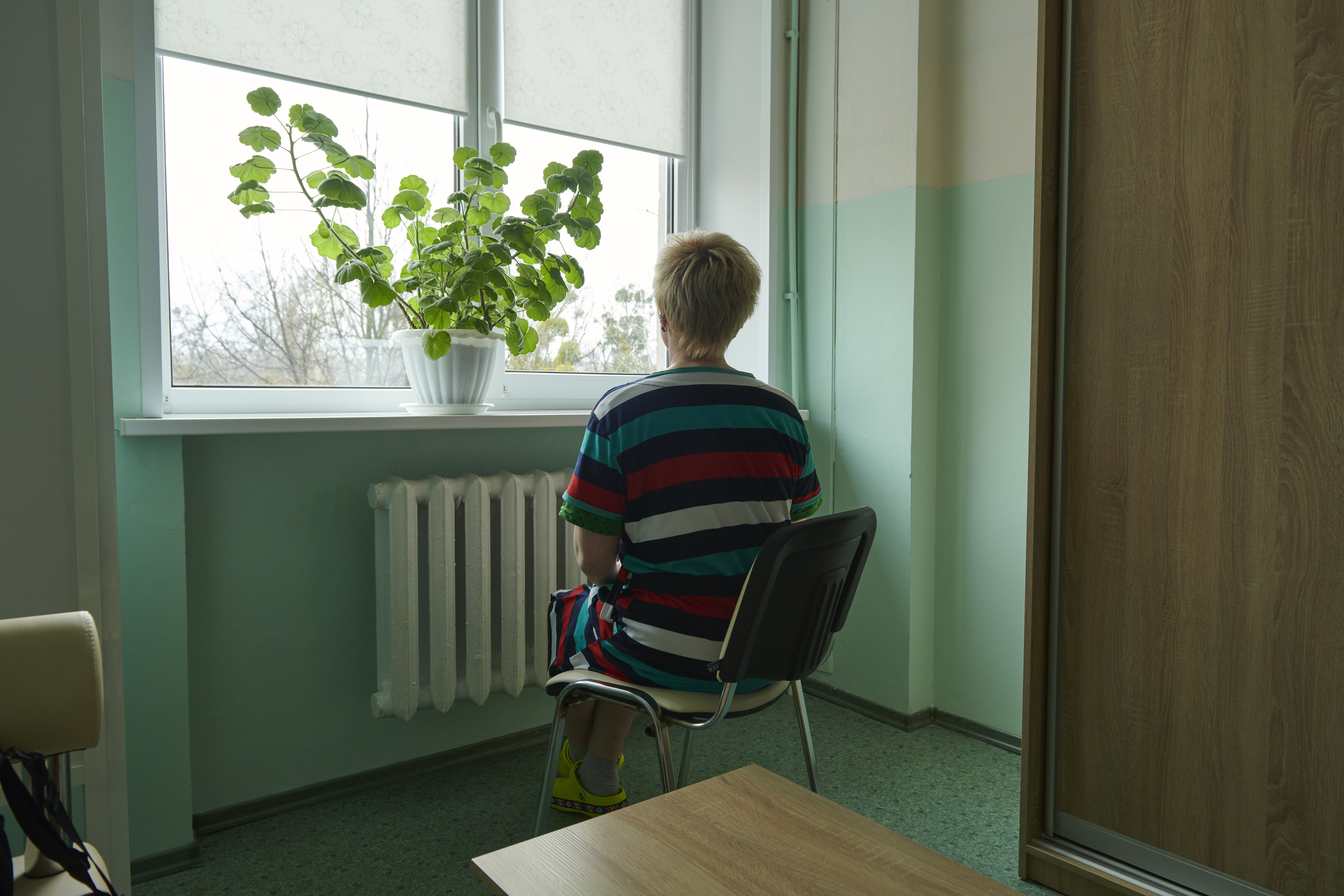 A Ukrainian women sits in a chair with her back to the camera facing the window. There is a green plant on the window ledge, and the room is painted a light green. 