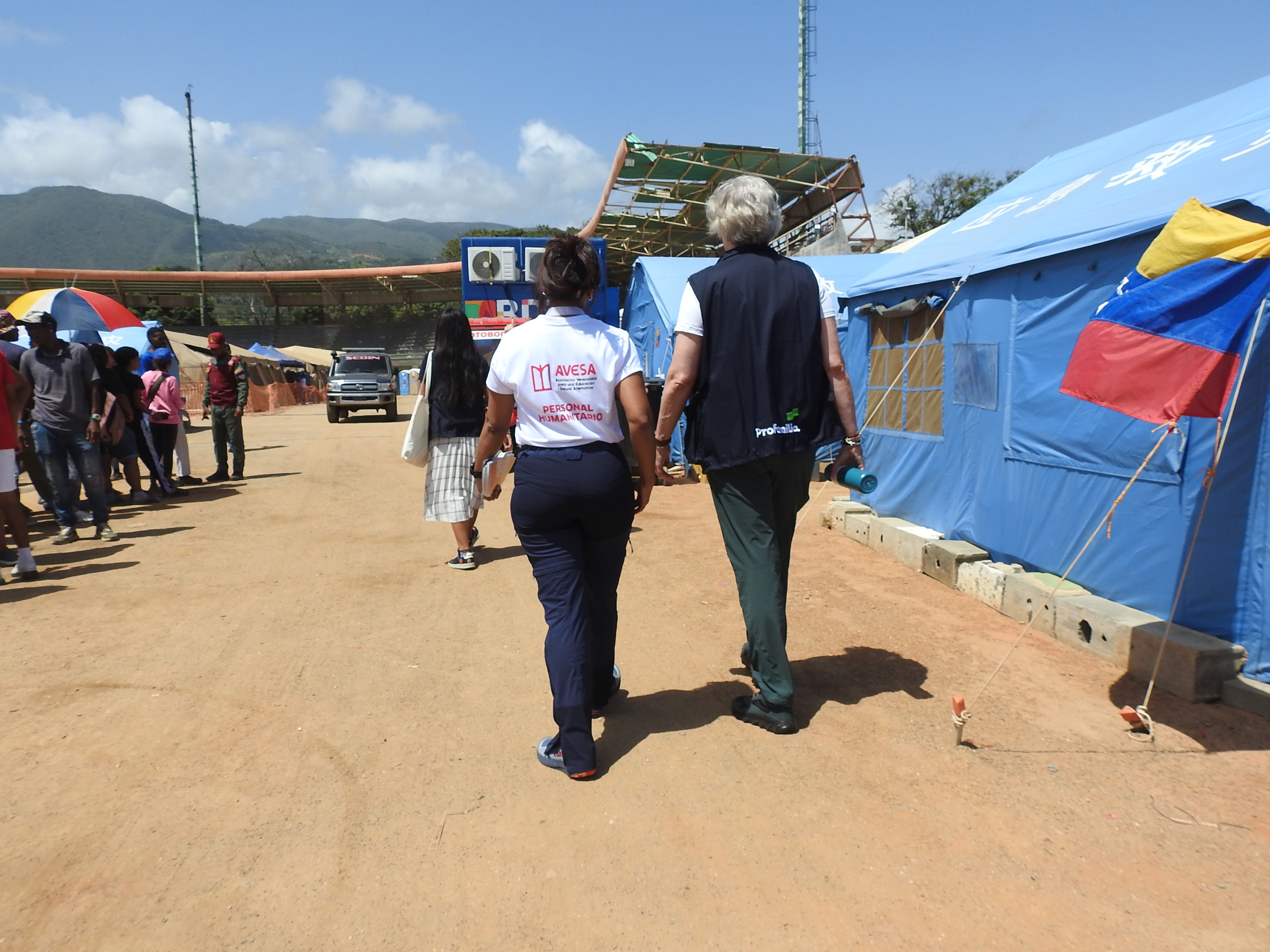 Two women walk along the dusty floor of an open air football stadium, filled with tents. One has the Venezuelan flag on it.