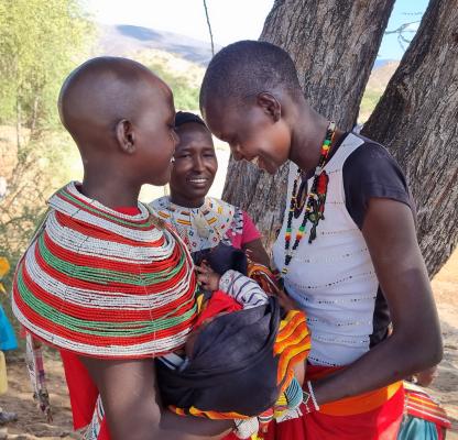 Three women stand over a baby in traditional dress in Kenya. They stand with their heads close together smiling. 