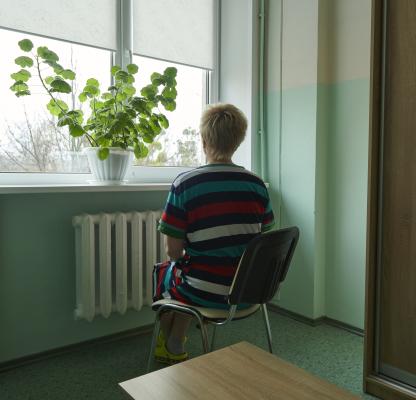 A Ukrainian women sits in a chair with her back to the camera facing the window. There is a green plant on the window ledge, and the room is painted a light green. 