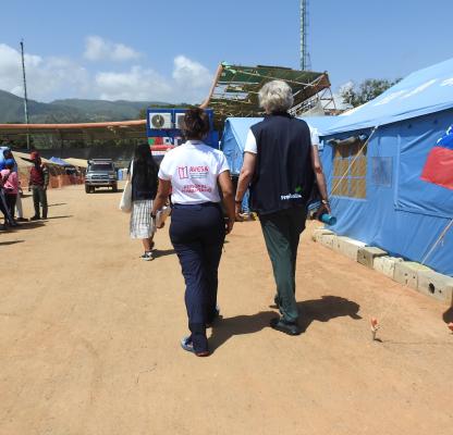 Two women walk along the dusty floor of an open air football stadium, filled with tents. One has the Venezuelan flag on it.