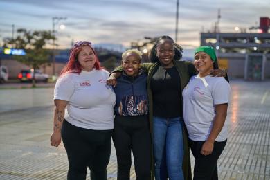 Four women stand together facing the camera with their arms around each other. 
