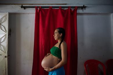 Colombia - I pregnant woman stands side on to the camera holding her bump. They are wearing a green top and blue shorts. 
