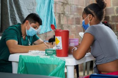 A woman in a grey t-shirt sits across a table from a health worker. Both are wearing face masks