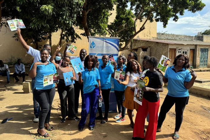 A group of peer educators gather in blue t-shirts waving leaflets and stood in front of a banner