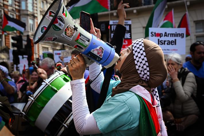 A woman stands shouting into a megaphone. She is draped in a Palestinian flag, and behind her are others holding up banner saying Stop Arming Isreal with more Palestinian flags. 