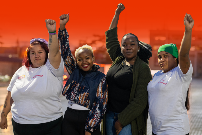 Four women stand in a row holding there fists in the air on a street with the IPPF fire red background colour