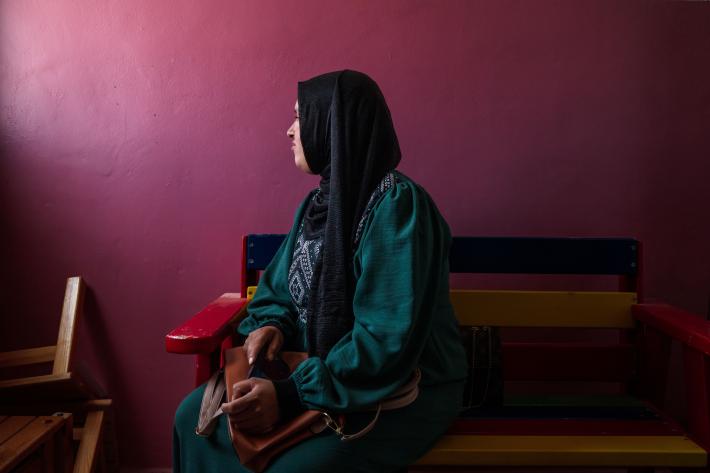  women in the West Bank sits on a bench, with her head turned towards the left. She is sat against a dark purple background. 