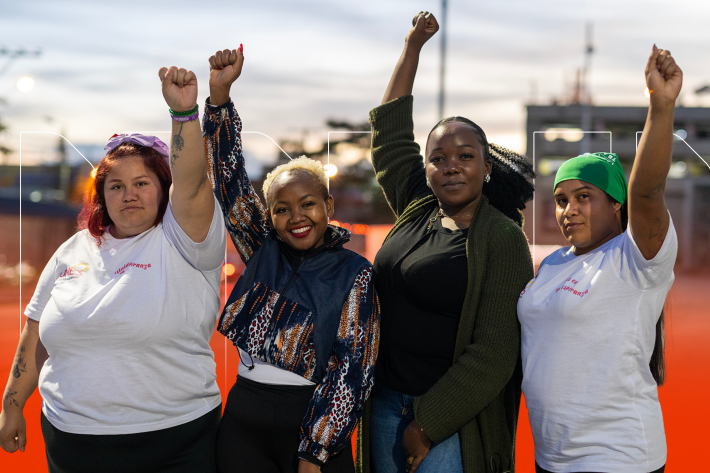 Four women stand in a row with their first raised in the air. There is a cityscape in the background.