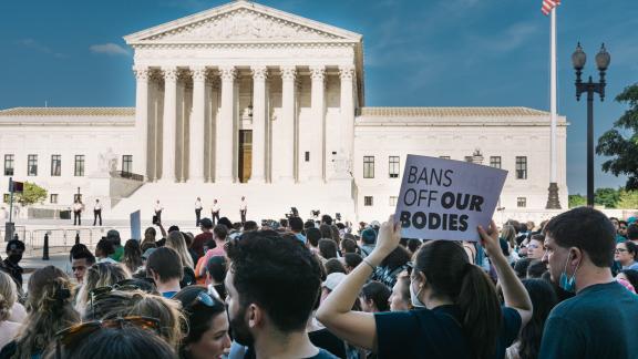 The US Supreme Court building with many protestors outside. One visible sign reads "Ban off our bodies"