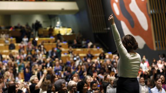 A women is shown speaking to a large audience. 