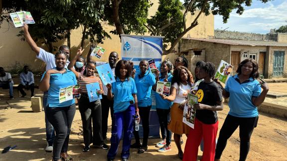 A group of peer educators gather in blue t-shirts waving leaflets and stood in front of a banner