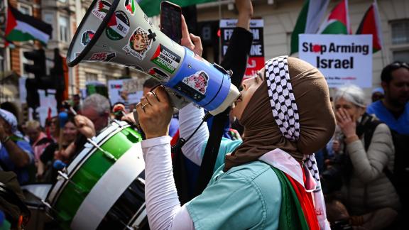 A woman stands shouting into a megaphone. She is draped in a Palestinian flag, and behind her are others holding up banner saying Stop Arming Isreal with more Palestinian flags. 