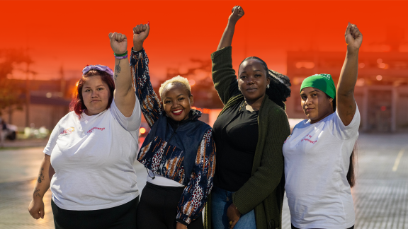 Four women stand in a row holding there fists in the air on a street with the IPPF fire red background colour