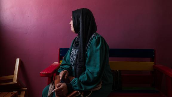  women in the West Bank sits on a bench, with her head turned towards the left. She is sat against a dark purple background. 