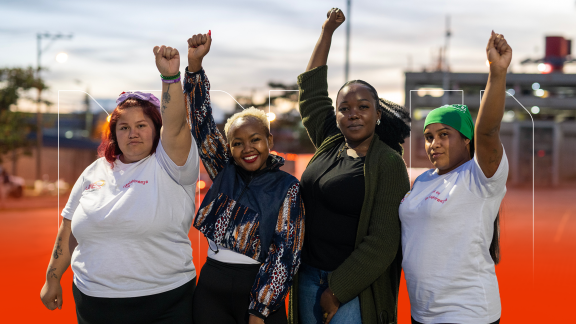Four women stand in a row with their first raised in the air. There is a cityscape in the background.